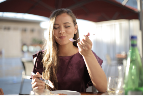 A woman eating at an outside cafe, smiling, wearing a maroon top.