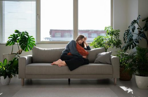A female sitting on a grey couch in an office with plants and an open window.