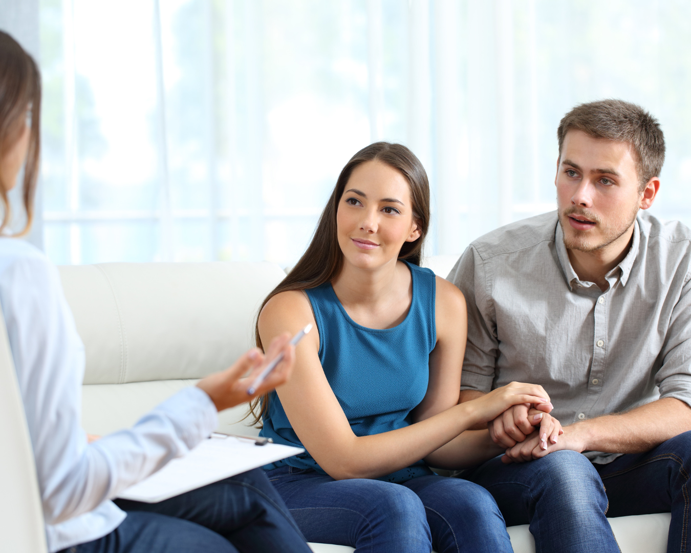Male/Female couple sitting on a couch, female is wearing a blue tank top and jeans, male is wearing a grey button up long sleeve shirt and jeans, they are holding hands, talking with a female sitting in a chair wearing a white long sleeve shirt holding a pen and notepad.