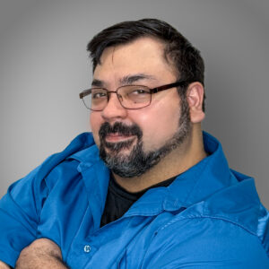 Man with dark hair and glasses and beard, in a blue shirt with arms crossed.